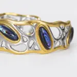 Close-up of an ONDA LENTA openwork cuff bracelet with iridescent oval abalone shell inlays, gold-tone borders and sculptural silver-tone metalwork.
