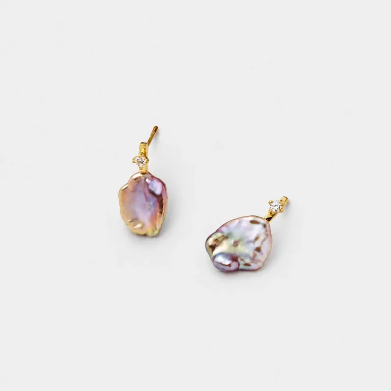 Angled detail view showing the iridescent pink and lavender glow of the natural flat baroque pearl studs with a sparkling prong-set stone accents.