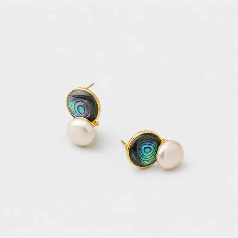 Angled detail view showing the shifting blue-green light of the circular abalone shell and white pearl stud earrings finished with polished 18K gold plated rims.