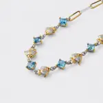 An angled close-up view showcasing the alternating square-cut blue topaz stones and two-tone textured fan charms with small red accent stones at the hinge details on the handcrafted 18k gold plated sterling silver bracelet.