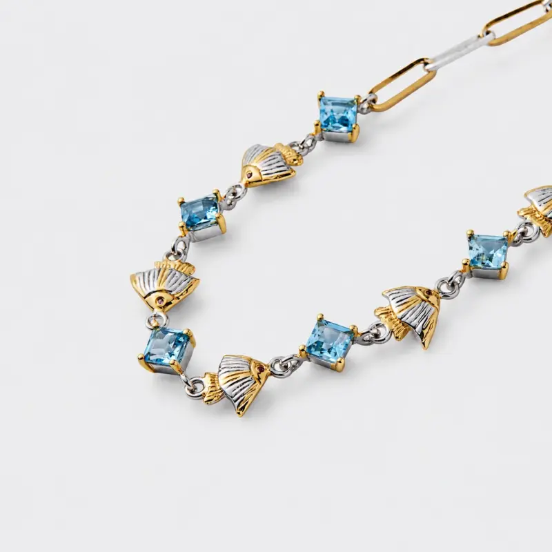 An angled close-up view showcasing the alternating square-cut blue topaz stones and two-tone textured fan charms with small red accent stones at the hinge details on the handcrafted 18k gold plated sterling silver bracelet.