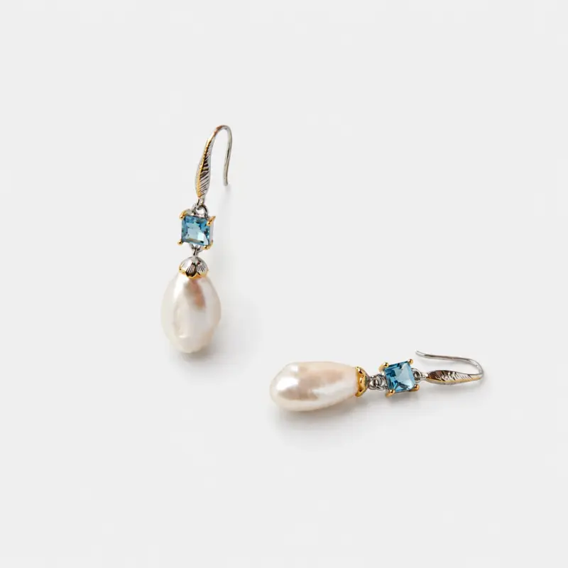 Close-up detailed view of the square-cut blue topaz stones and shell-shaped two-tone metal elements above naturally irregular baroque pearl drop earrings crafted in 18K gold plated sterling silver