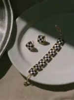 ONDA LENTA luxury 18K gold-plated jewelry set showcasing a square-link checkerboard pattern bracelet and matching stud earrings, elegantly arranged on a white ceramic plate under dramatic cinematic shadows.