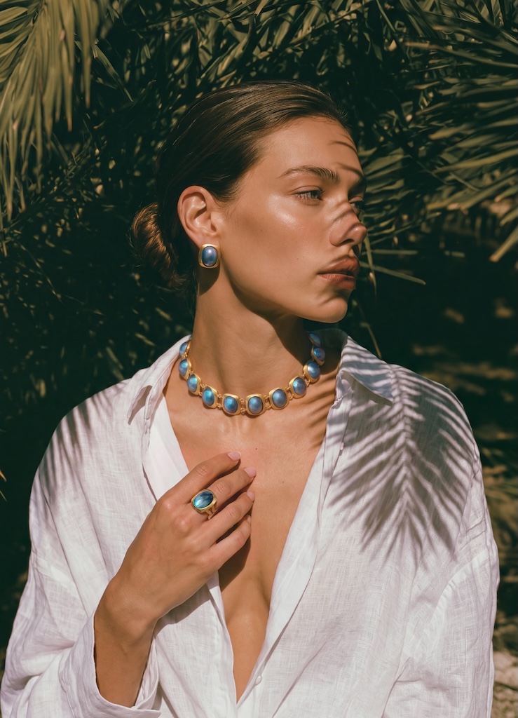 Model wearing ONDA LENTA luxury 18K gold-plated jewelry set, featuring a blue iridescent statement necklace, matching stud earrings, and a large cocktail ring under natural sunlight with palm leaf shadows.